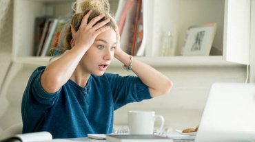 Portrait of an attractive woman at the table with cup and laptop, book, notebook on it, grabbing her head. Bookshelf at the background, concept photo